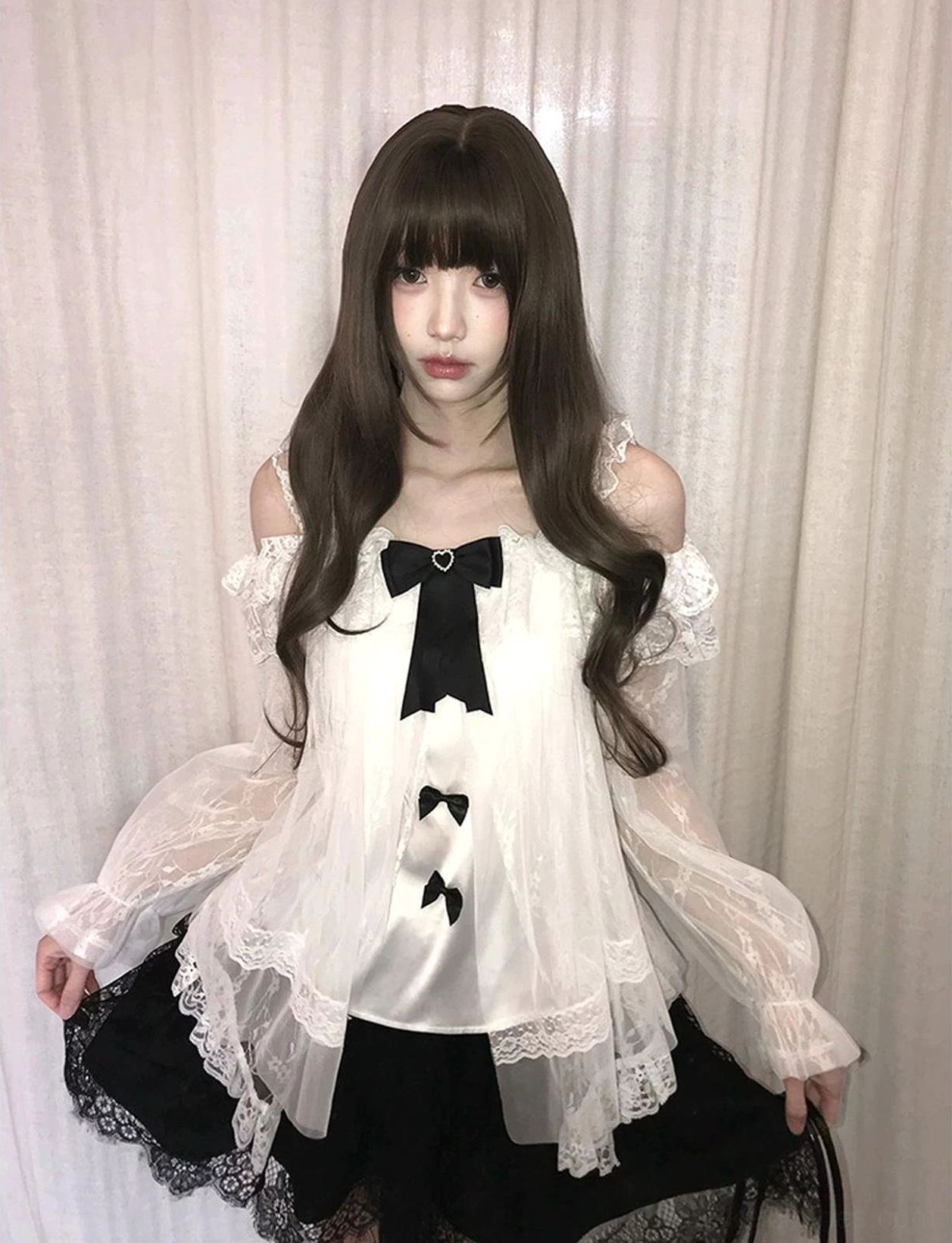 Half-body front view of black-and-white off-shoulder sheer lace blouse with long ruffled sleeves, detachable bows, and delicate lace hem trim worn indoors against curtain background