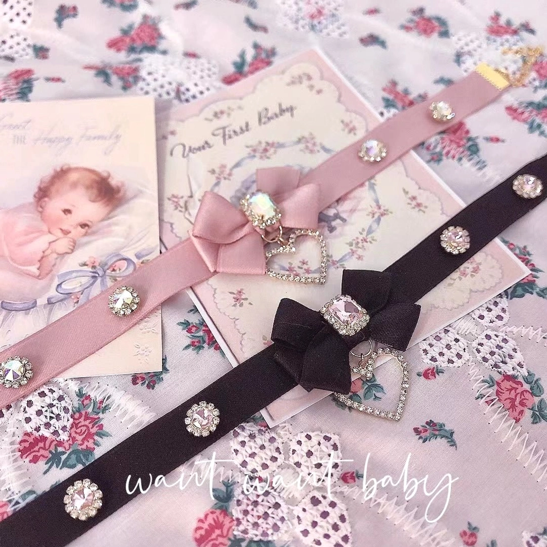Both pink and black bow rhinestone choker necklaces displayed on vintage floral card backdrop with gold-tone clasps and heart pendants visible