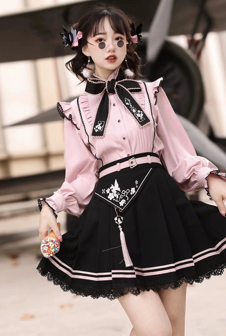 Pink and black Jirai Kei two-piece outfit with ruffled blouse and pleated skirt, full front view on young female model with vintage airplane background
