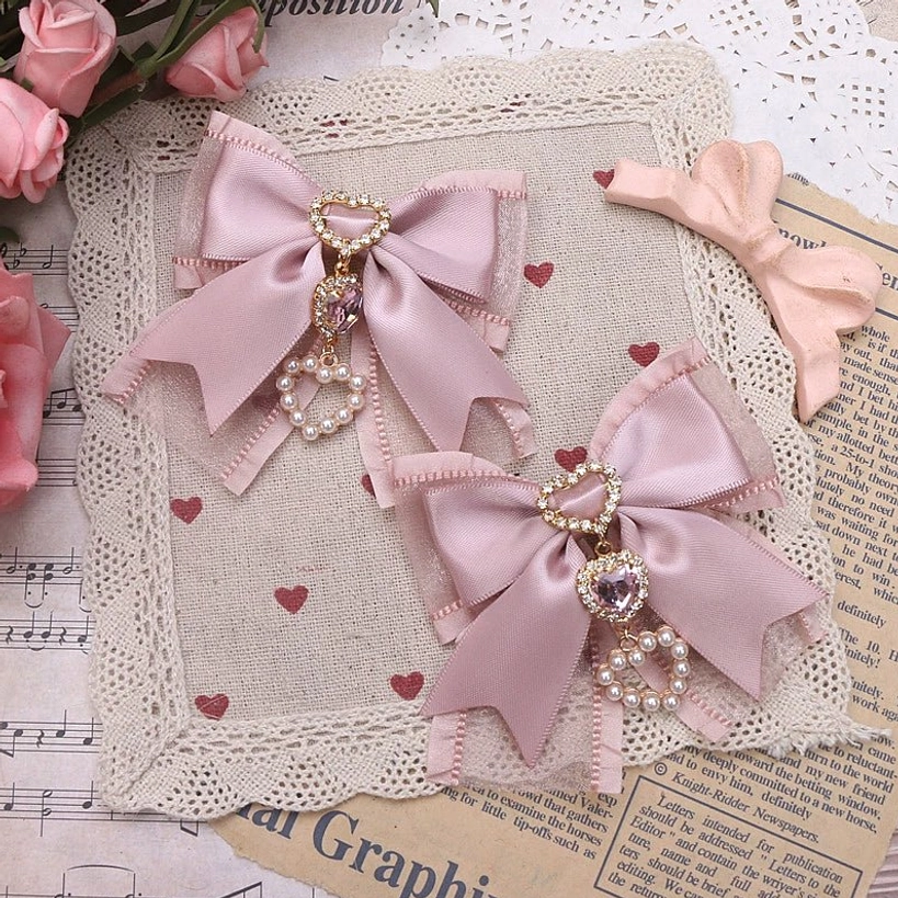 Two dusty pink bow hair clip styles with heart rhinestone pendants displayed flat on vintage lace fabric background