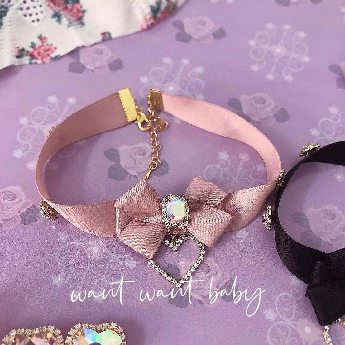 Close-up detail of pink satin bow rhinestone choker necklace featuring central square gem and heart-shaped rhinestone pendant on purple floral background