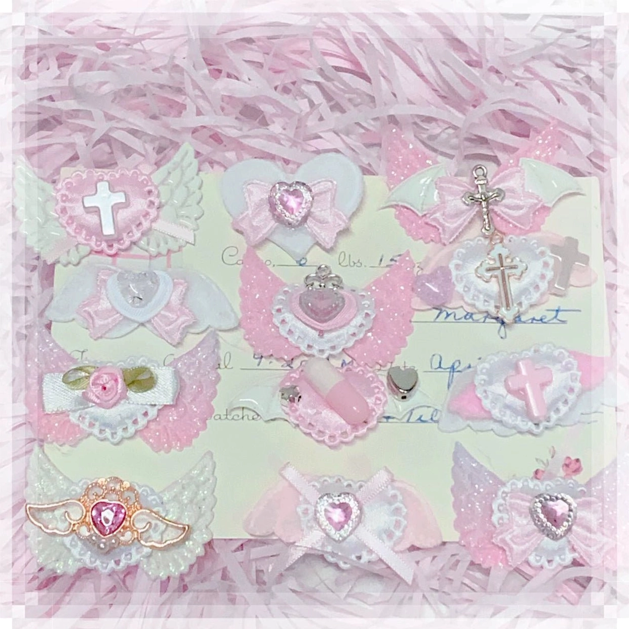Assorted pink jirai kei hair clips top-down flat lay on pink shredded paper with display card showing cross heart rhinestone and iridescent wing decorations