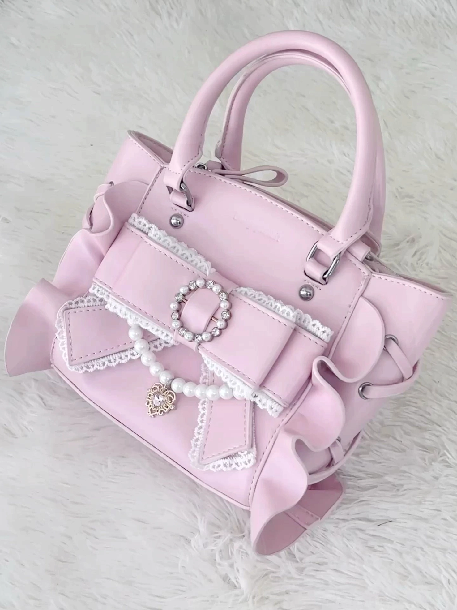 Pink Jirai Kei square handbag flat lay overhead view featuring rhinestone bow pendant, lace trim, pearl chain, and ruffled side details on white fluffy carpet background