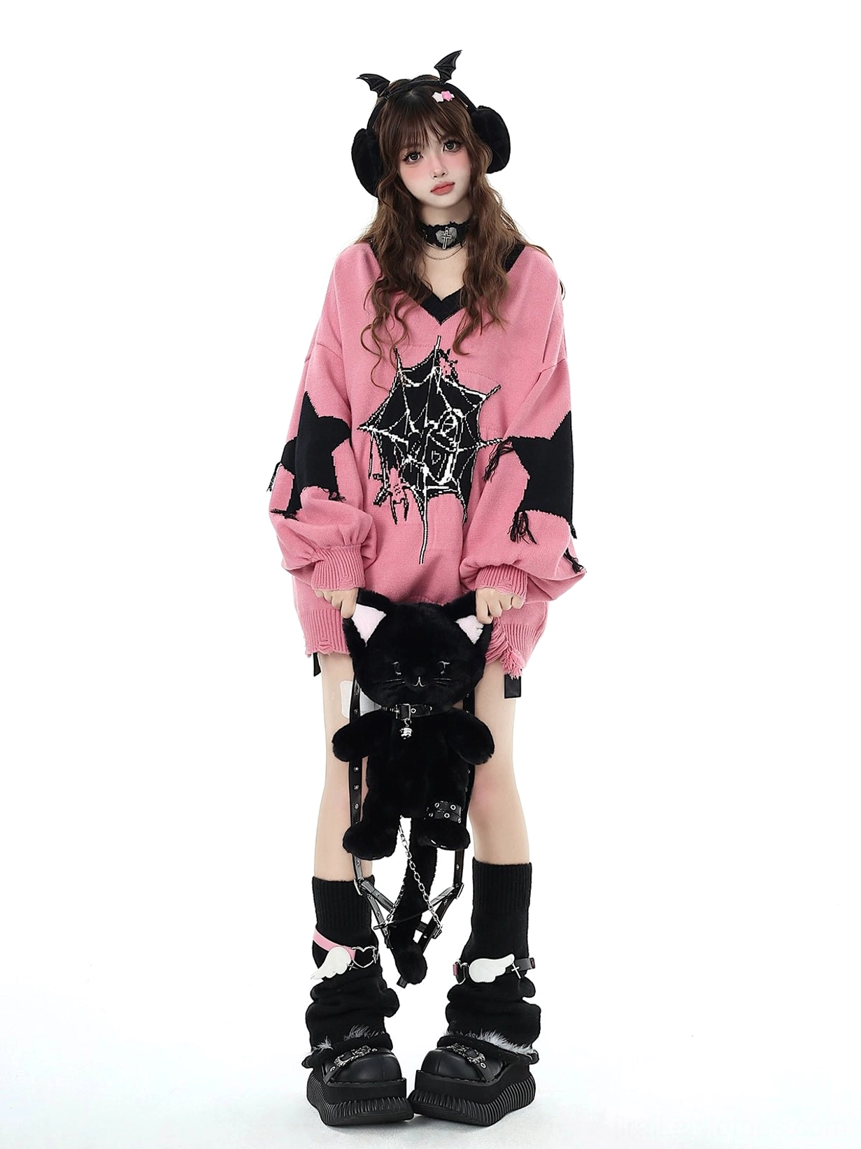 Pink Jirai Kei oversized knit sweater with star shoulder prints and distressed hem worn by model with devil horn earmuffs holding plush cat ornament