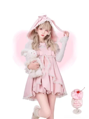 Pink ryousangata lolita dress with detachable bunny ear hood worn by blonde model in three-quarter kneeling pose, holding plush rabbit with pink dessert backdrop and ruffled lace hem detail visible
