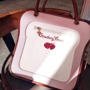 Pink two-tone Toast Ita Bag top-down close-up showing zipper front pocket and embroidered strawberry charm on wooden chair