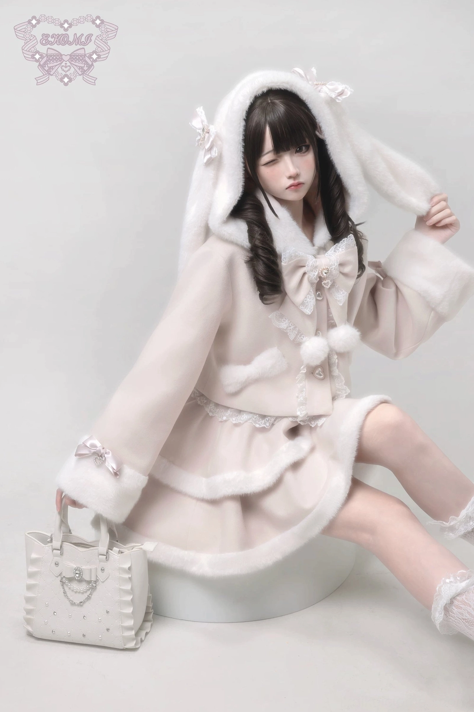 Ryousangata rabbit-ear hooded lolita coat in beige with bunny hood up, sitting pose side view holding matching studded handbag, fluffy fur details and lace accents visible