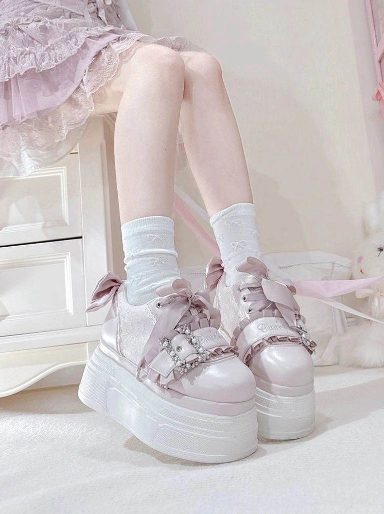 Light pink Jirai Kei platform shoes worn on feet with pink lace outfit and white socks, front three-quarter view showing rhinestone buckle and satin bow decoration