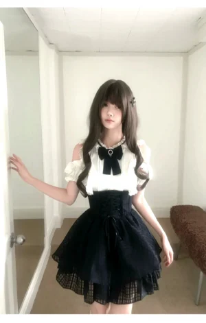 Seiso Kei cold shoulder blouse and high-waist black plaid skirt set worn by model in indoor full-length mirror setting, featuring white ruffled top with puff sleeves and detachable rhinestone bow tie