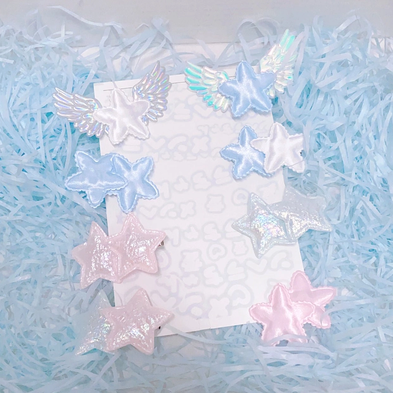 Star wing hair clips collection assorted flat lay with blue shredded paper featuring pink blue and silver star shapes with angel wing embellishments