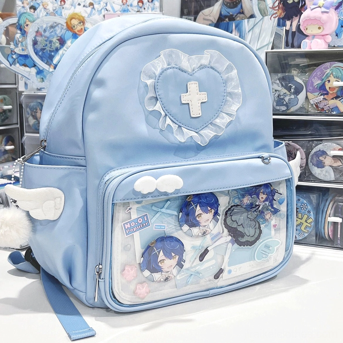 Kawaii blue itabackpack with transparent collection pocket displayed on desktop with anime figures, featuring angel wing side accents and heart lace cross ornament
