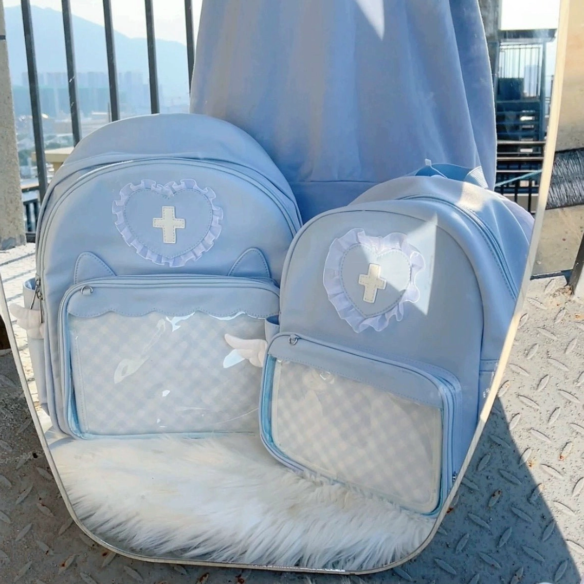 Two pastel blue itabackpacks front upper view on rooftop with metal floor and fur mat background, featuring heart lace cross badges, clear display pockets with grid patterns and angel wing side details