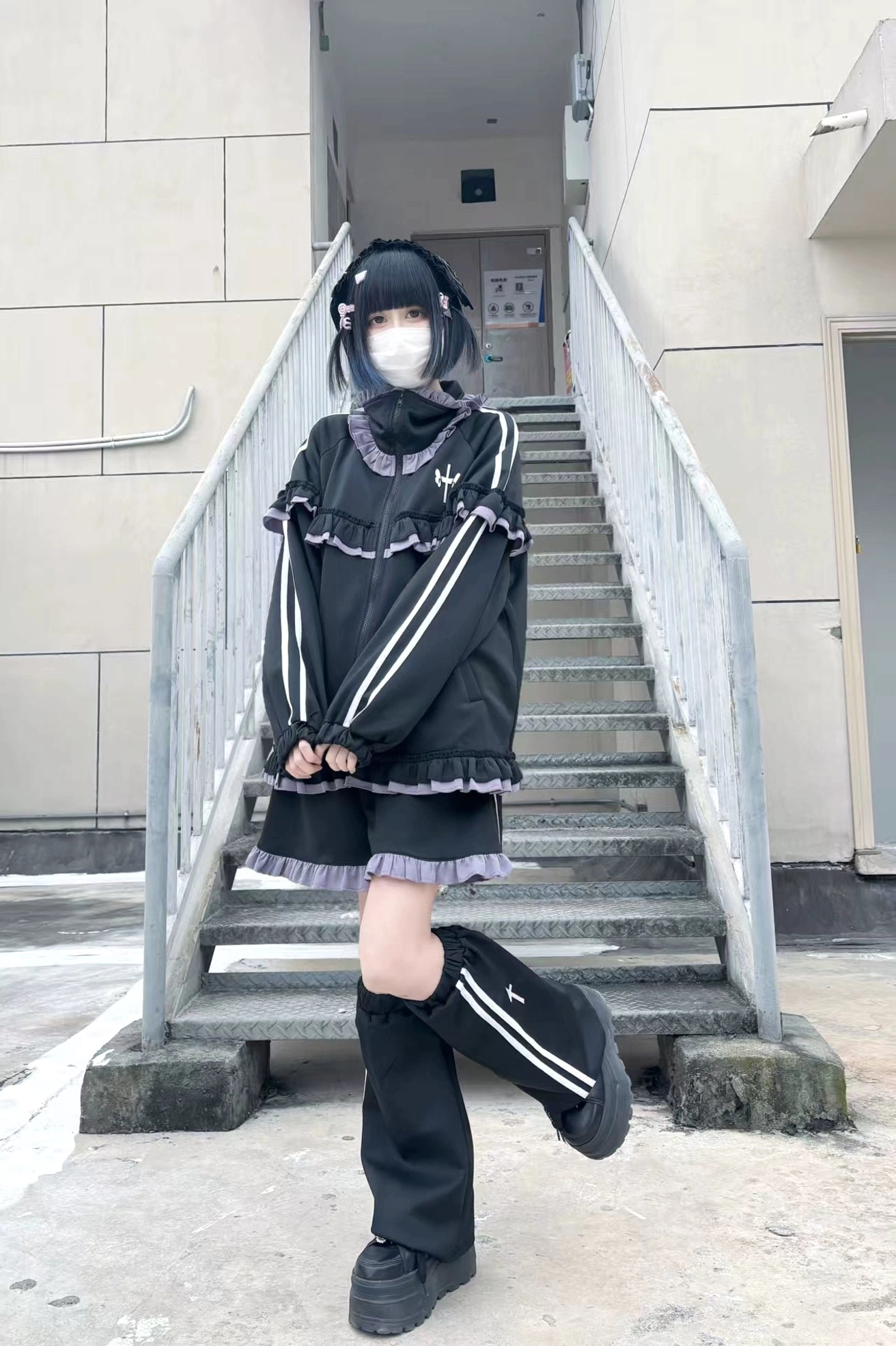 Black-purple ruffle Tenshi Kaiwai tracksuit with striped leg warmers, frill trim shorts and loose zip-up jacket on apartment stairway
