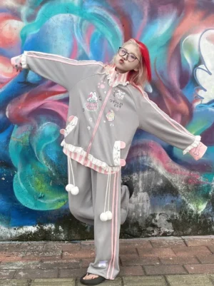 Grey Tenshi Kaiwai kawaii coat and pants ensemble with ruffle details and pink trim worn by model with red hair against graffiti wall outdoor setting angel style tracksuit