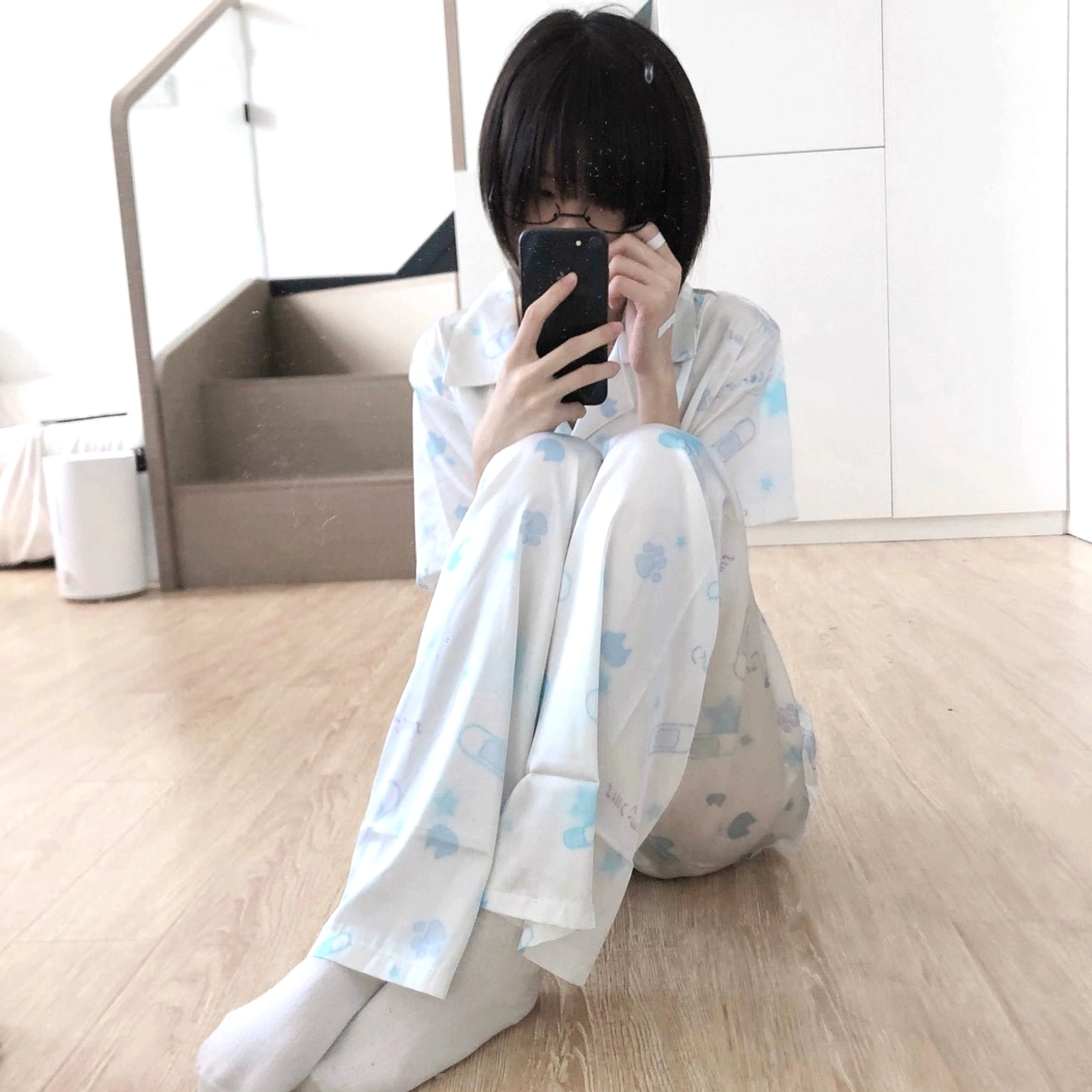 Female model wearing light blue Tenshi Kaiwai cat print long-sleeve pajama top with pants and white socks, front selfie in home wooden floor bedroom setting