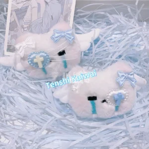 Tenshi Kaiwai plush crying kitty hair clip set flat lay featuring pastel pink and blue variants with angel wings cross-heart patches and bow accents on light blue shredded filler background with anime postcard