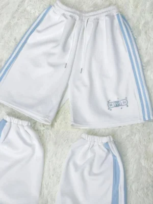 Tenshi Kaiwai white bi-color shorts with light blue vertical stripes and OK bandage embroidery on left leg, flat lay display on furry surface showing adjustable drawstring waist and front logo detail
