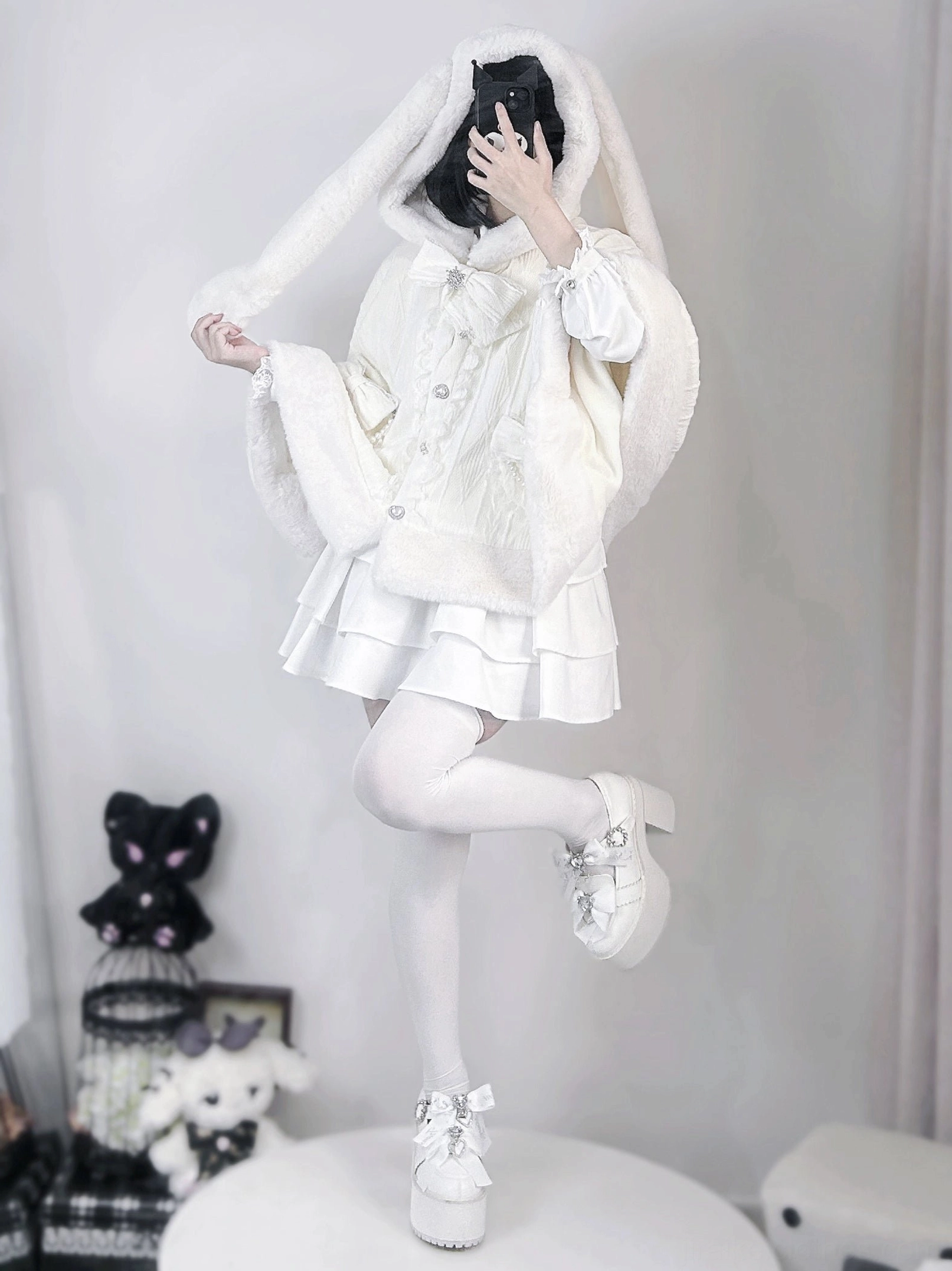 Side profile mirror selfie of white rabbit ear cape coat on model with lifted leg pose, featuring plush rabbit ears and white platform loafers