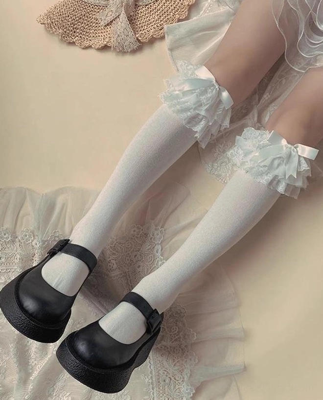 White Jirai Kei ruffled bow socks with asymmetric ruffled satin bow trim pair of legs black Mary Jane shoes lace mat background side overhead view