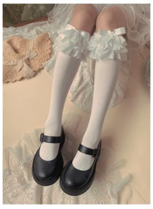 White Jirai Kei lace frills mid-calf socks with satin ribbon bows paired with black Mary Jane shoes on vintage lace mat background overhead front view