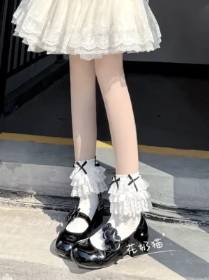 White frilly Jirai Kei mid-calf socks with black bows worn with white mini skirt and outdoor concrete staircase background, front lower leg styling view