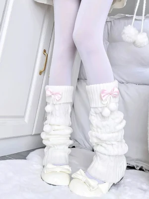 White knitted Jirai Kei leg warmers with pink ribbon bow and fluffy pom-poms, worn by model in indoor home setting, featuring ruched knit texture and feminine styling details