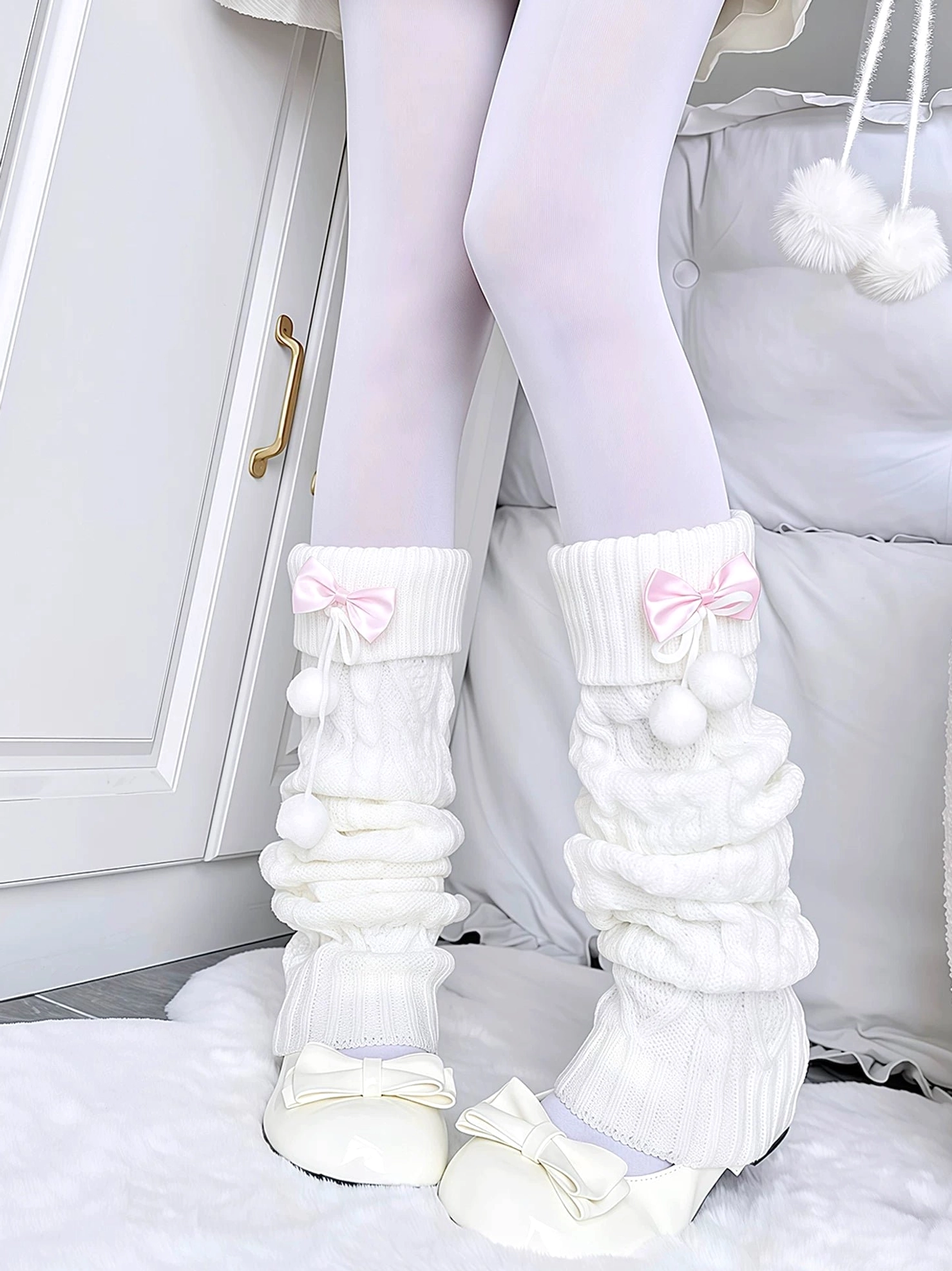 White knitted Jirai Kei leg warmers with pink ribbon bow and fluffy pom-poms, worn by model in indoor home setting, featuring ruched knit texture and feminine styling details