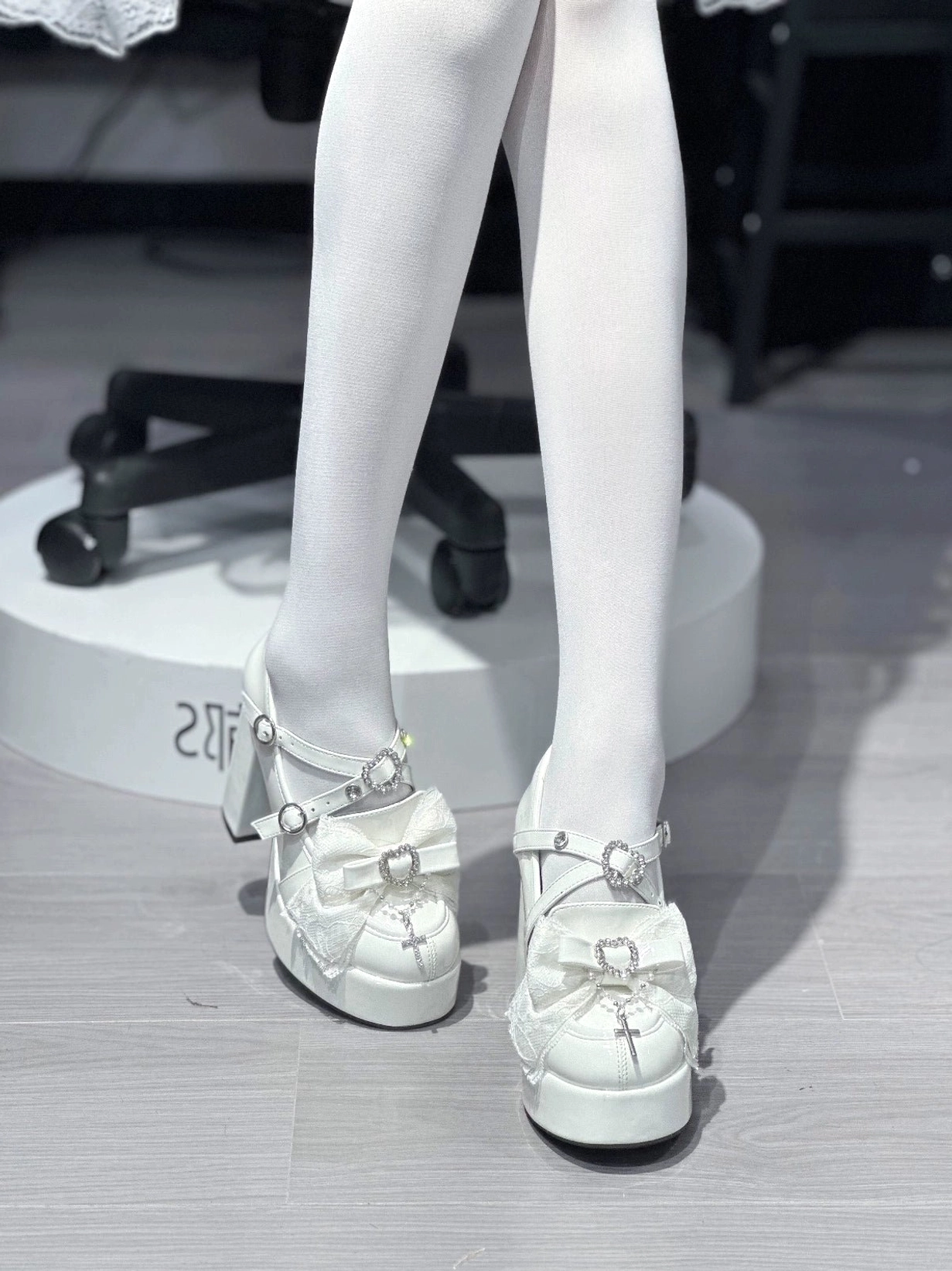 Detail close-up of white Jirai Kei platform heels highlighting heart-shaped rhinestone embellishments, lace bow decoration on vamp, and patent leather shine