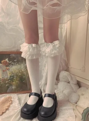 White Jirai Kei ruffled lace mid-calf socks with double-layer frills worn with black Mary Jane shoes against vintage indoor decor backdrop straight front view