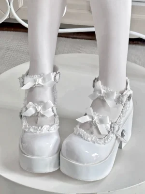 White patent leather Jirai Kei platform shoes with ornamental cross charm and heart-shaped rhinestone embellishments, front overhead view displaying chunky platform sole, dual ankle straps, satin bow accents, and delicate lace edging